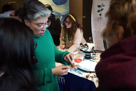 Una mujer con pelo canoso y gafas muestra un objeto natural a un grupo de personas interesadas en la ciencia.