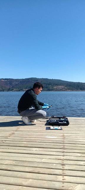 Un hombre está manipulando un dispositivo en un muelle junto a un lago en un día soleado.