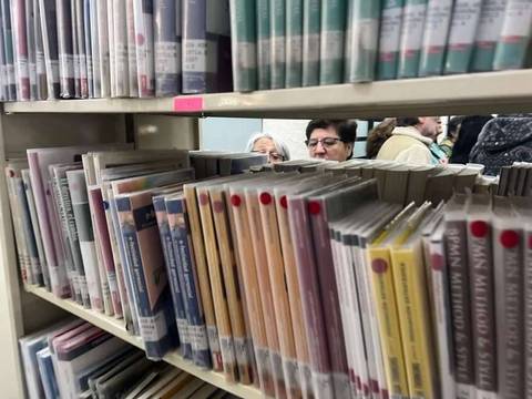 En una biblioteca, se pueden ver libros y dos personas al fondo observando.