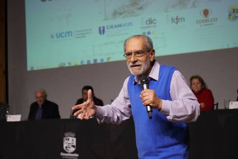 Un hombre mayor en un suéter azul habla apasionadamente con un micrófono en la mano en un evento académico.