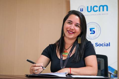 Una mujer sonriente firma documentos con un fondo institucional.