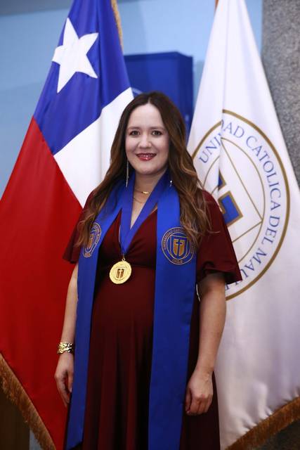 Una mujer embarazada con vestido rojo y medalla sonríe en un evento académico junto a banderas chilenas y del universidad.