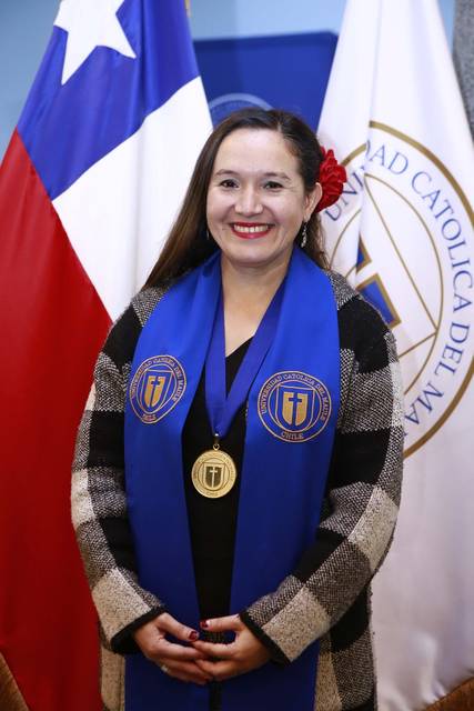 Una mujer sonriente con una banda azul y una medalla, posando frente a banderas chilena y de la universidad.