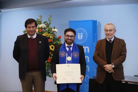 Un joven celebra su graduación con un diploma en manos, acompañado de dos personas en un evento institucional.