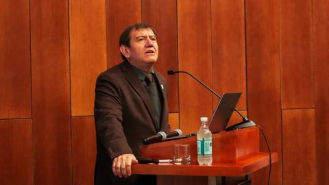 Un hombre está dando una charla frente a un público en una sala con paneles de madera.