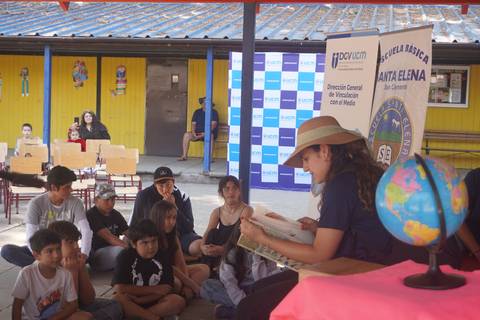 Un grupo de niños escucha atentamente a una persona que lee un libro bajo un techado en una escuela.