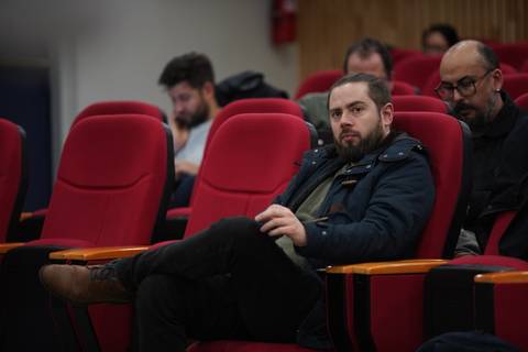 Un grupo de personas sentadas en un auditorio con sillas rojas, algunos parecen atentos a lo que ocurre adelante.
