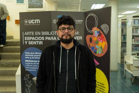 Un joven con barba y gafas posando frente a un muro colorido en una biblioteca.