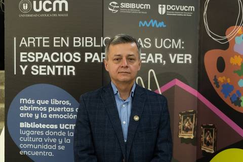 Un hombre de pie frente a un fondo que promociona actividades artísticas en la Universidad Católica del Maule.