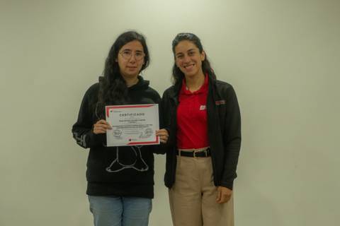 Una joven muestra su certificado junto a una mujer en un ambiente formal.