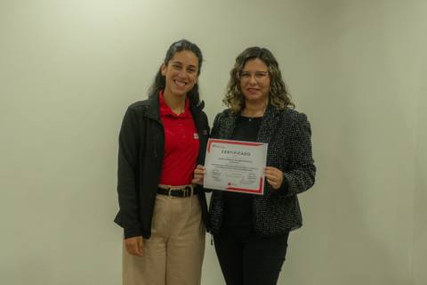 Dos mujeres posan juntas mostrando un certificado en un ambiente formal.