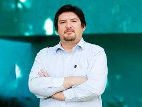 Un hombre con brazos cruzados posando frente a un fondo verde.