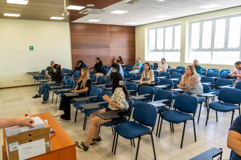 Un grupo de estudiantes atentos en un aula con sillas azules y mucha luz natural.
