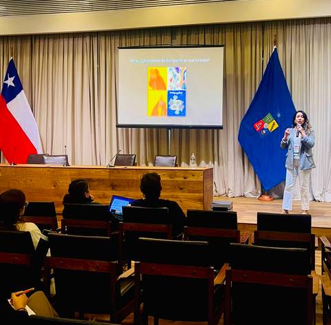 Una expositora presenta en un seminario mientras se ven banderas chilenas de fondo.