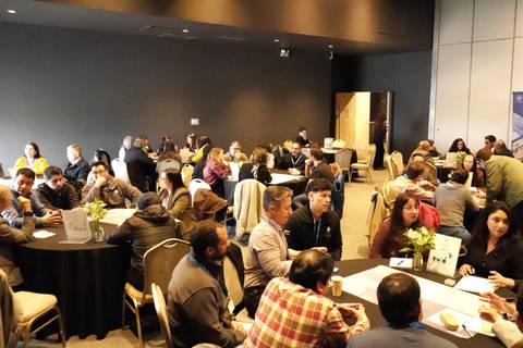 En la imagen se puede ver un evento con varias personas conversando en grupos alrededor de mesas en una sala amplia.