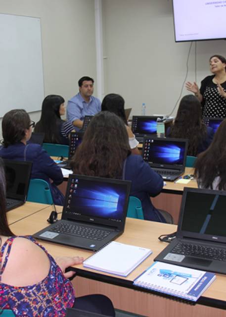 Una clase con alumnos en sus computadoras, mientras un profesor y una profesora dan una explicación.