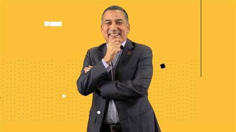 Un hombre sonriente de pie, vestido con un traje, posando con confianza frente a un fondo amarillo.