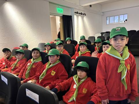 Un grupo de niños con chaquetas rojas y gorros verdes, sentados en un auditorio.