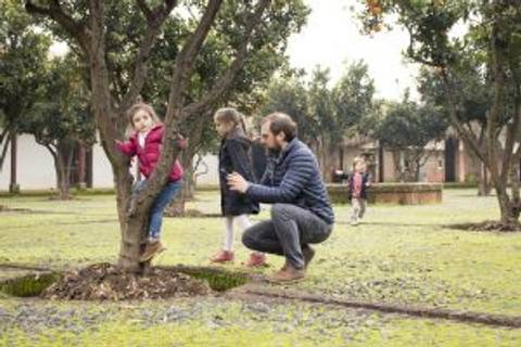 Un padre juega con sus hijas en un parque, donde una de ellas se trepa a un árbol mientras las otras observan.