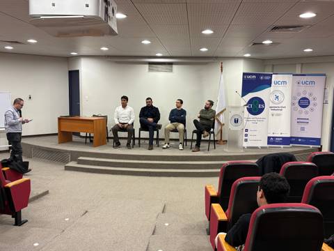 Un panel de discusión en una sala de conferencias con varios participantes sentados y un moderador de pie.