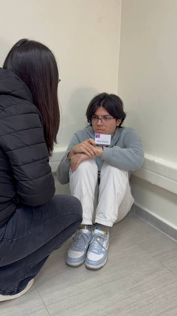 En la imagen se ve a un joven sentado en una esquina con una expresión de preocupación mientras una persona lo observa de cerca.