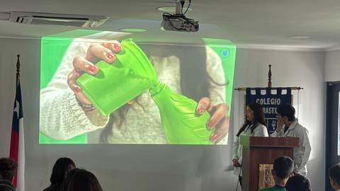Un par de estudiantes presentan un experimento mientras sostienen un recipiente verde frente a una proyección.