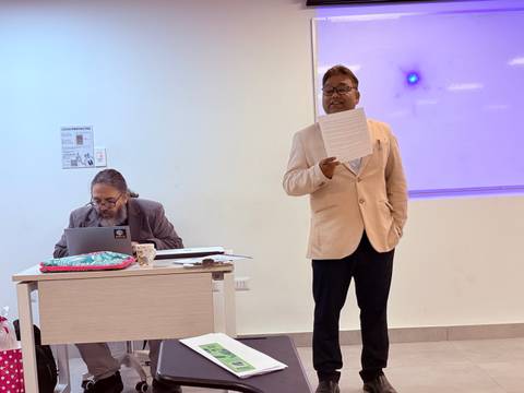 Un hombre habla frente a un grupo en una sala de clases mientras otro trabaja en su computador.
