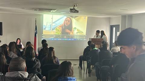 Una presentación en un salón con una pantalla mostrando a una persona, mientras otra persona habla frente a un público.