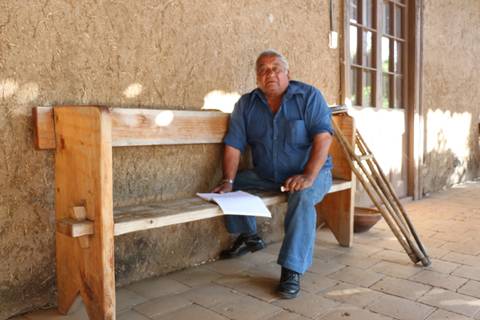 Un hombre sentado en un banco de madera frente a una pared de adobe.