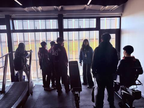 Un grupo de jóvenes se encuentra en un salón mirando hacia el exterior a través de unas grandes ventanas.