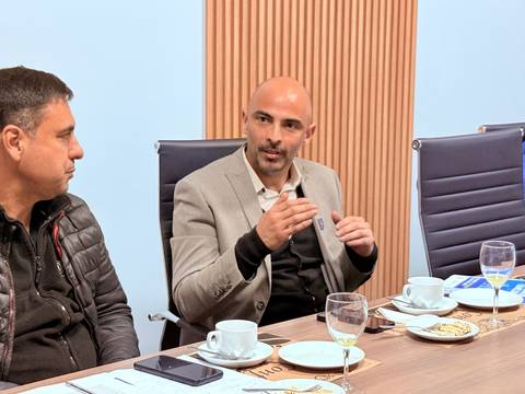 En una reunión formal, un hombre con chaqueta habla mientras su compañero lo escucha atentamente.