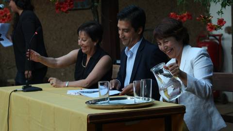 Tres personas sentadas en una mesa durante un evento, una de ellas sirviendo agua.
