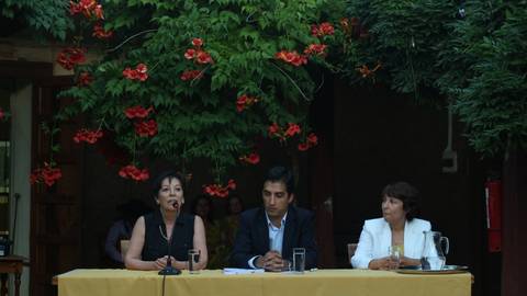 Tres personas participan en una mesa de discusión rodeada de plantas y flores rojas.