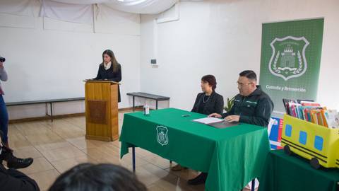 Una presentación formal con un ponente en un podio y dos personas sentadas en una mesa frente a él.