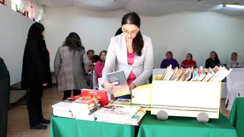 Una mujer revisa libros en una mesa mientras otras personas están sentadas al fondo en un evento.