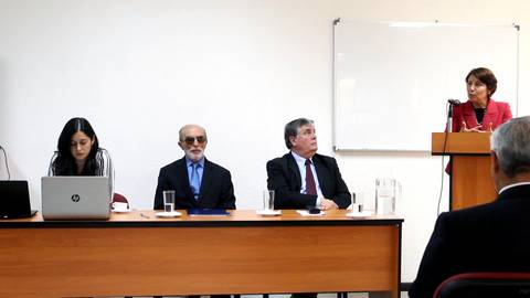 Una reunión formal con varias personas sentadas frente a una mesa, mientras una mujer está de pie hablando.