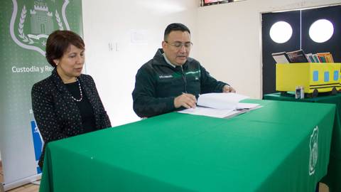 Una mujer y un hombre se encuentran sentados frente a una mesa verde, el hombre revisa documentos mientras la mujer observa.