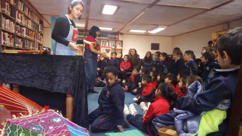 Un grupo de niños observa una representación en una biblioteca, mientras dos presentadoras interactúan con ellos.
