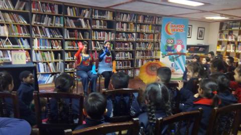 Una actuación con marionetas en una biblioteca, donde dos personas entretienen a un grupo de niños sentados frente a ellas.
