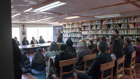 Una reunión comunitaria en una biblioteca con un gran grupo de personas sentadas y una mujer hablando frente a ellas.