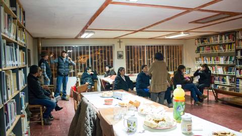 Una reunión en un salón con estanterías llenas de libros donde algunas personas conversan y otras están sentadas.