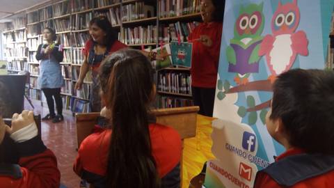 Un grupo de niños escucha atentamente una narración en una biblioteca, mientras dos narradoras presentan un cuento ilustrado.