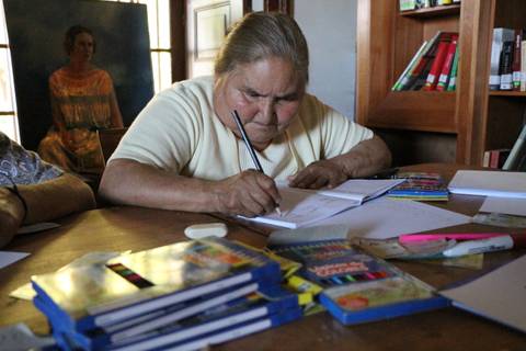 Una mujer mayor está escribiendo en un cuaderno en una mesa llena de materiales de escritura.