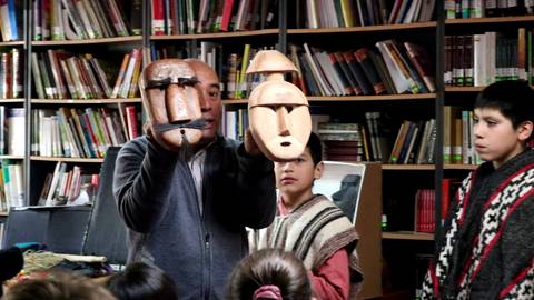 Un hombre muestra tres máscaras a un grupo de niños en una biblioteca.