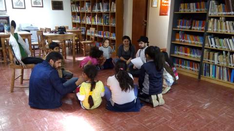 Un grupo de niños y adultos se reúne en una biblioteca para realizar una actividad en conjunto.
