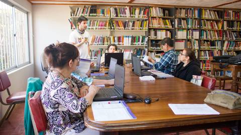 Un grupo de personas trabaja en un aula con computadoras mientras un facilitador explica algo frente a ellas.