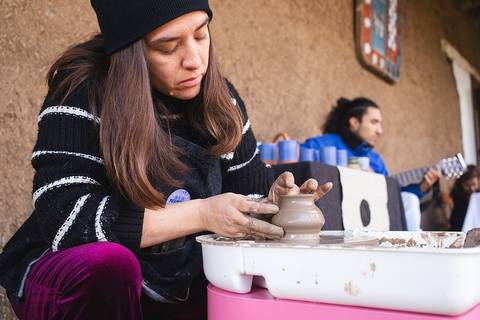 Una mujer modela arcilla en un torno de alfarería mientras un hombre toca la guitarra de fondo.