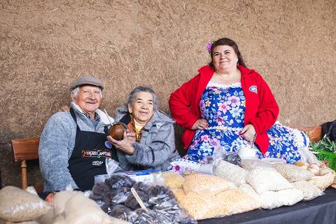 Tres personas sonrientes posan junto a una mesa llena de productos locales en un mercado.