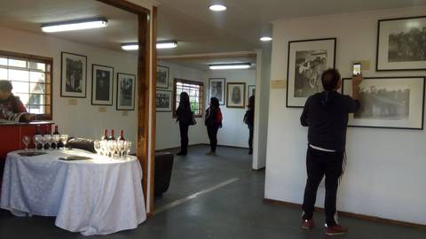 Una galería de arte con varias personas observando fotografías enmarcadas y una mesa con copas de vino.