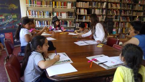Un grupo de niños y adultos se encuentran en una biblioteca, participando en actividades creativas alrededor de una mesa.